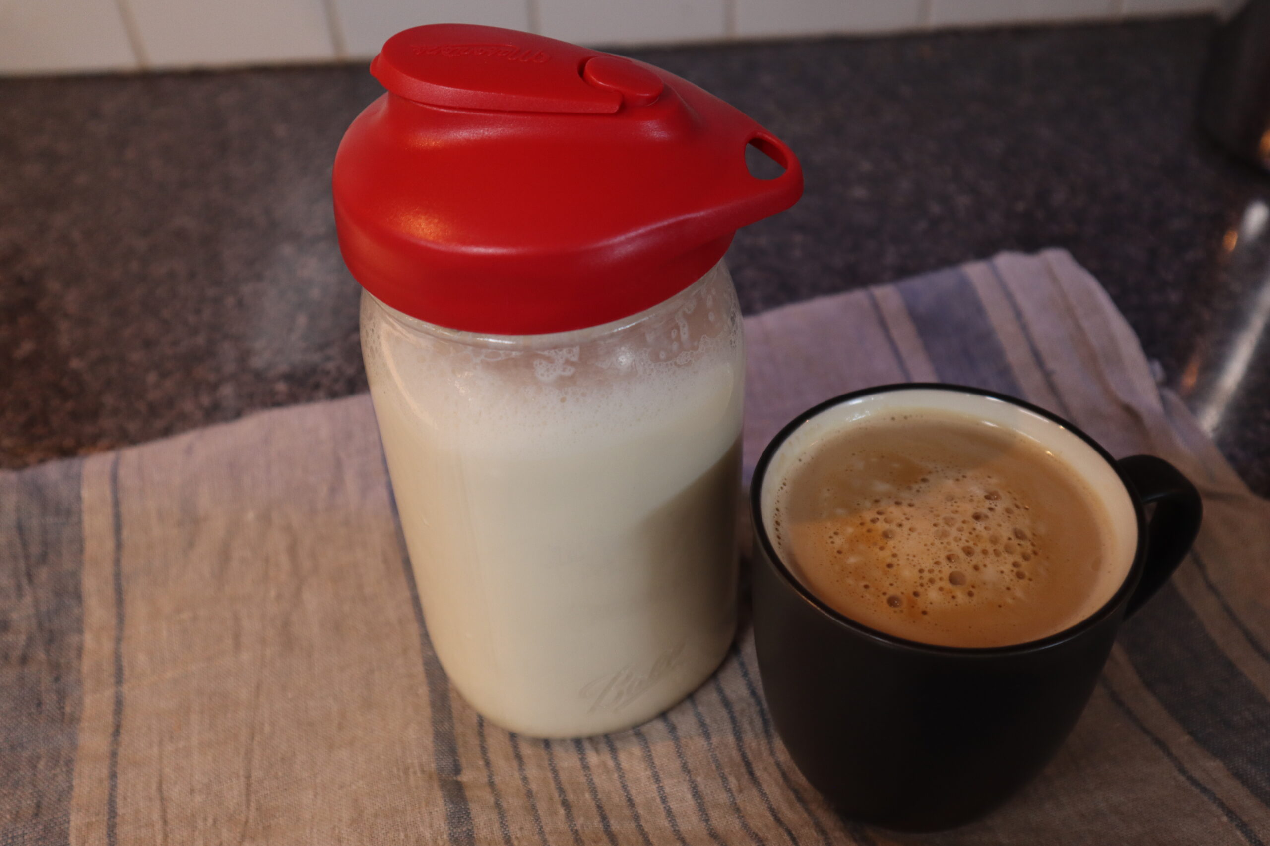 Homemade coffee creamer in a mason jar with a red lid, besides a cup of coffee, both on top of a gray and blue linen striped kitchen towel