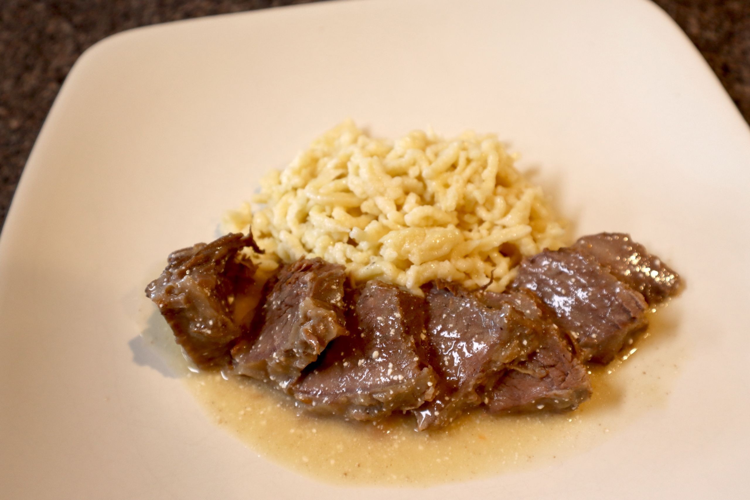 Sauerbraten sliced and topped with gravy, served with spaetzle on a white plate